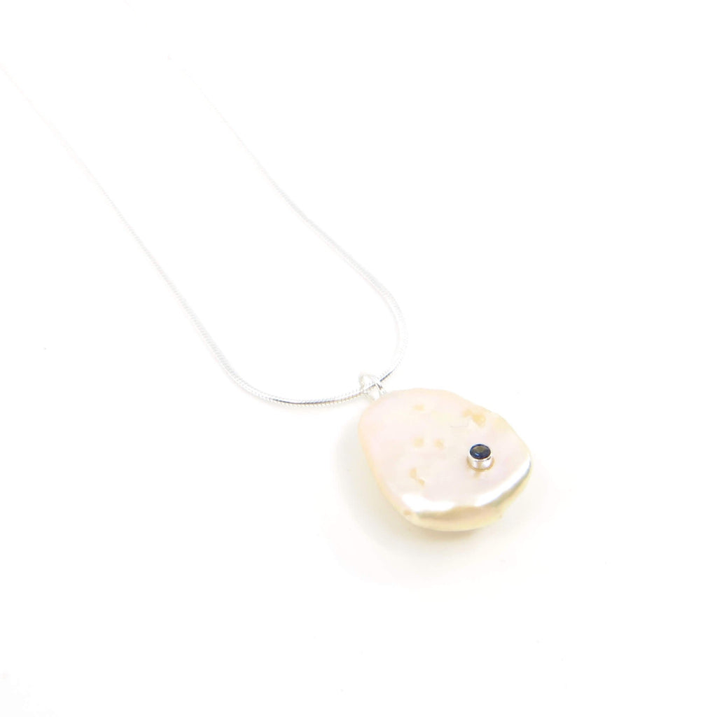 A peachy champagne keshi pearl embedded with a sterling silver tube set 2mm very dark blue sapphire. Pendant is suspended from a 16" sterling silver snake chain.