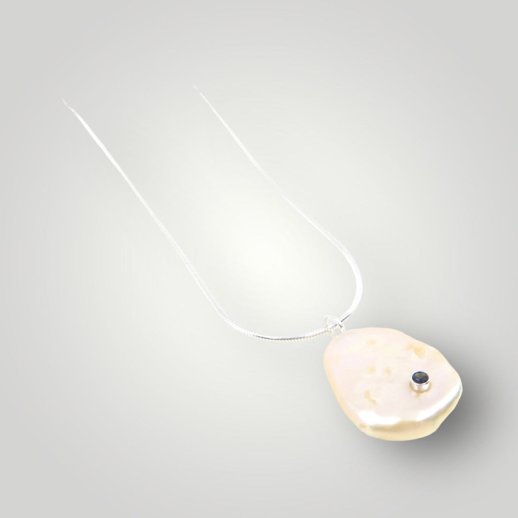 A peachy champagne keshi pearl embedded with a sterling silver tube set 2mm very dark blue sapphire. Pendant is suspended from a 16" sterling silver snake chain.